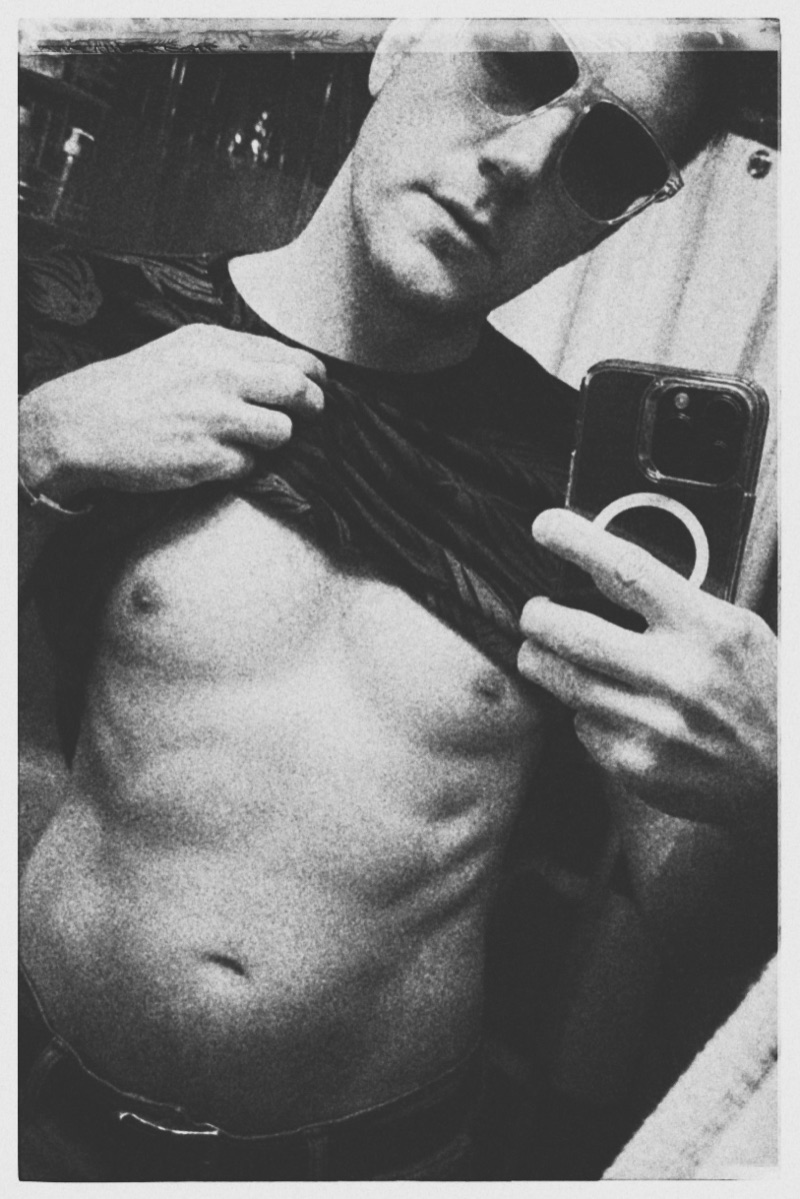 A black-and-white mirror photograph, phone raised, shirt lifted, sunglasses on.