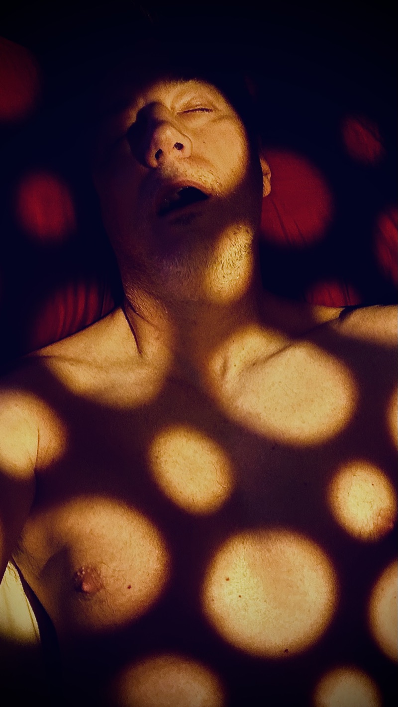 Fugdale lying back with eyes closed under yellow and blue light, scattered coloured spots falling across the frame.